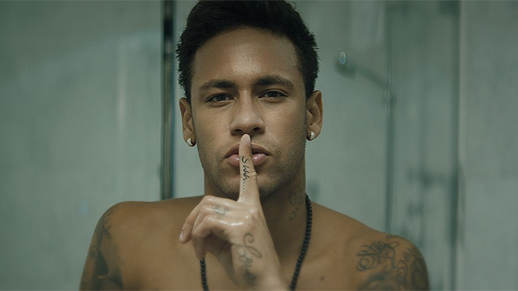 a shirtless man with his finger at his lips in a quieting gesture