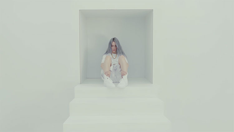 a young white woman with long hair wearing all white and sitting at the top of a set of white stairs in a white room