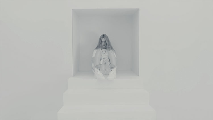 a young white woman with long hair wearing all white and sitting at the top of a set of white stairs in a white room