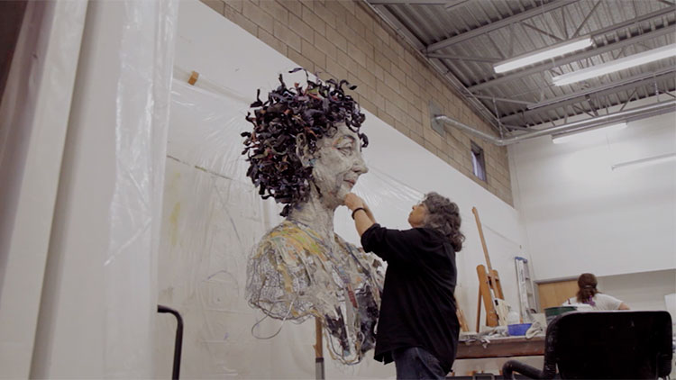an older white woman artist working on a very large female bust sculpture